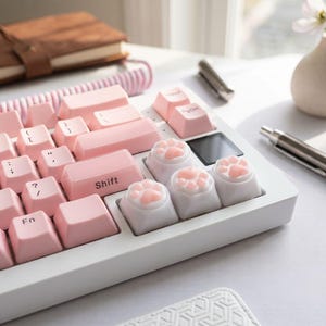 Puede incluir: Un teclado mecánico blanco con teclas de color rosa claro con letras negras y tres teclas blancas especiales con forma de patas de gato en relieve con almohadillas de color rosa suave. El teclado incluye un cable rosa en espiral y una pequeña pantalla negra.