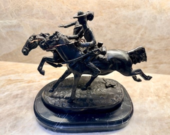 May include: A dark bronze sculpture depicting two riders on horseback in a dynamic pose. The sculpture is mounted on a black marble base. The horses are in mid-stride, conveying a sense of motion and action.