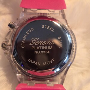 May include: A clear wristwatch with a pink silicone band. The watch face is black with silver text that reads "Geneva Platinum" and "Japan Movt". The watch has a stainless steel back.