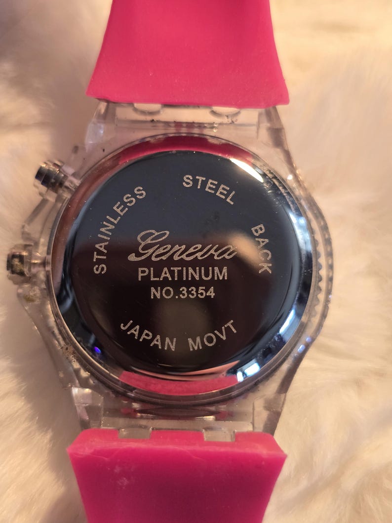 May include: A clear wristwatch with a pink silicone band. The watch face is black with silver text that reads "Geneva Platinum" and "Japan Movt". The watch has a stainless steel back.