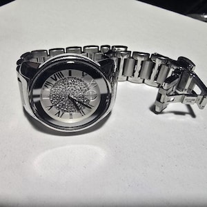 May include: A silver-toned wristwatch with a round face and a linked band. The watch face features Roman numerals and a central area covered in small, sparkling stones. The band is made of polished metal links.