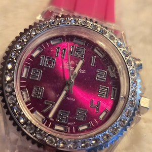 May include: A wristwatch with a pink face and a clear, rhinestone-studded bezel. The watch has a pink band and silver-colored hands and numerals. The watch face has the word "Geneva" printed on it.