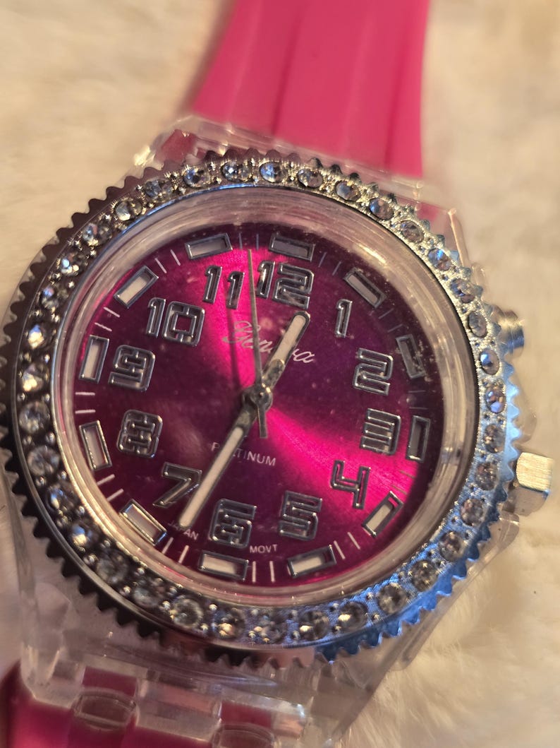 May include: A wristwatch with a pink face and a clear, rhinestone-studded bezel. The watch has a pink band and silver-colored hands and numerals. The watch face has the word "Geneva" printed on it.
