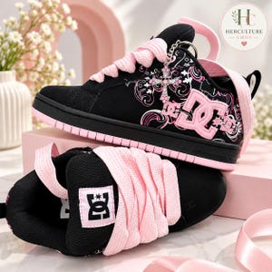 Puede incluir: Un par de zapatillas DC negras y rosas con cordones rosas gruesos, una base negra con patrones gráficos blancos y rosas, y un dije de cruz plateada unido a los ojales. La lengüeta muestra un parche con el logo DC blanco sobre fondo negro.