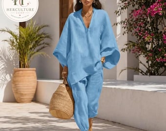 May include: A light blue linen two-piece outfit. The top has a V-neck and wide sleeves, paired with matching cropped pants. The outfit is worn with gold-colored sandals and a woven tote bag. The HerCulture Shop logo is in the upper left corner.