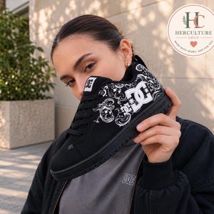 Puede incluir: Una zapatilla negra DC con un patrón floral tipo cachemira en blanco en el lateral, un logotipo DC blanco, cordones negros y suela negra. El calzado cuenta con un cuello acolchado y un pequeño parche con el logotipo DC en la lengüeta.