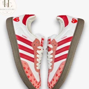 Op de afbeelding: Een paar witte Adidas Samba sneakers met rode strepen, witte veters en een bruine rubberen zool. De schoenen zijn versierd met rode hartjes op de hiel en een roze hartjespatroon op de neus. Het woord Samba staat in het rood op de zijkant.