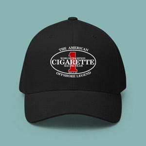 Könnte beinhalten: Eine schwarze Baseballkappe mit einer weißen ovalen Grafik, die den Text THE AMERICAN, WORLD CHAMPION, CIGARETTE RACING TEAM, OFFSHORE LEGEND und MADE IN USA enthält, zentriert um eine große rote Nummer 1. Die Kappe hat eine strukturierte Krone.