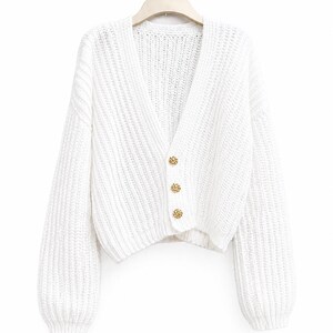 Op de afbeelding: Een witte, grofgebreide cardigan met een V-hals en lange mouwen. De trui heeft drie goudkleurige knopen aan de voorkant. Het kledingstuk wordt op een witte hanger tegen een witte achtergrond getoond.