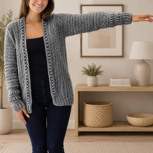 May include: A gray, open-front crochet cardigan with long sleeves. The cardigan has a textured stitch pattern and a ribbed cuff. The person is wearing a black top and dark blue jeans. The cardigan is a versatile layering piece.
