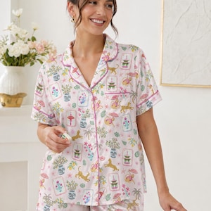 May include: Pink pajama set with a whimsical print featuring pagodas, tigers, and floral arrangements. The short-sleeved top and shorts are trimmed with pink piping. The model is holding a mahjong tile.