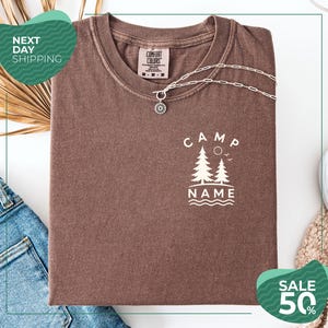 a brown t-shirt with a white design, a necklace, and some clothing items, including a pair of jeans, on a white background.