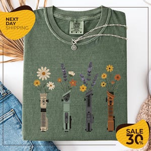 a green t-shirt with a graphic design of four different colored vases holding flowers, arranged in a row. The vases are depicted in various colors, including green, yellow, orange, and purple. The t-shirt is displayed on a white background, and there are yellow and black sale signs indicating a 30% discount.