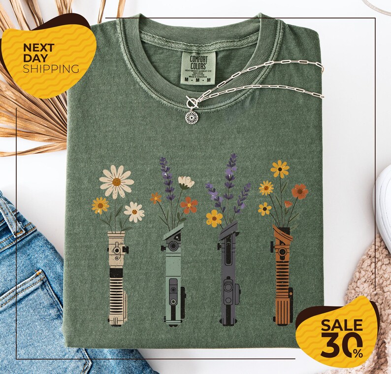 a green t-shirt with a graphic design of four different colored vases holding flowers, arranged in a row. The vases are depicted in various colors, including green, yellow, orange, and purple. The t-shirt is displayed on a white background, and there are yellow and black sale signs indicating a 30% discount.