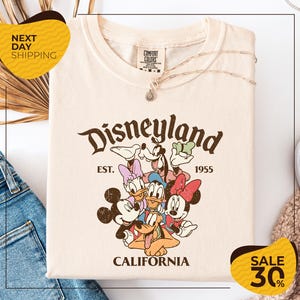 a white t-shirt with a Disney-themed graphic, including Mickey Mouse, Donald Duck, and Goofy, along with the text "Disneyland EST. 1955 CALIFORNIA". The t-shirt is displayed on a white background, and there are yellow and black sale signs indicating a 30% discount.