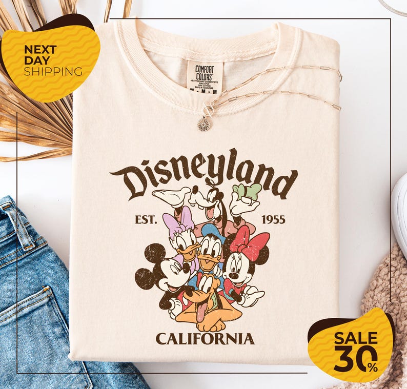 a white t-shirt with a Disney-themed graphic, including Mickey Mouse, Donald Duck, and Goofy, along with the text "Disneyland EST. 1955 CALIFORNIA". The t-shirt is displayed on a white background, and there are yellow and black sale signs indicating a 30% discount.