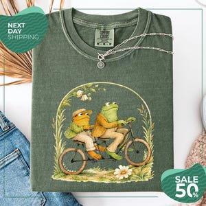 May include: An olive green t-shirt featuring a graphic illustration of two frogs riding a tandem bicycle. The frogs are wearing jackets and hats, surrounded by a floral archway. The shirt has a Comfort Colors label at the neck and a silver chain.