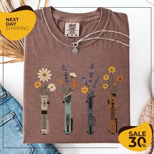 a brown t-shirt with a graphic design of four different colored vases holding flowers, arranged in a row. The vases are depicted in various colors, including green, gray, orange, and yellow. The design also includes a pendant necklace and a chain. The t-shirt is displayed on a white background, and there are yellow and black sale signs indicating a 30% discount.