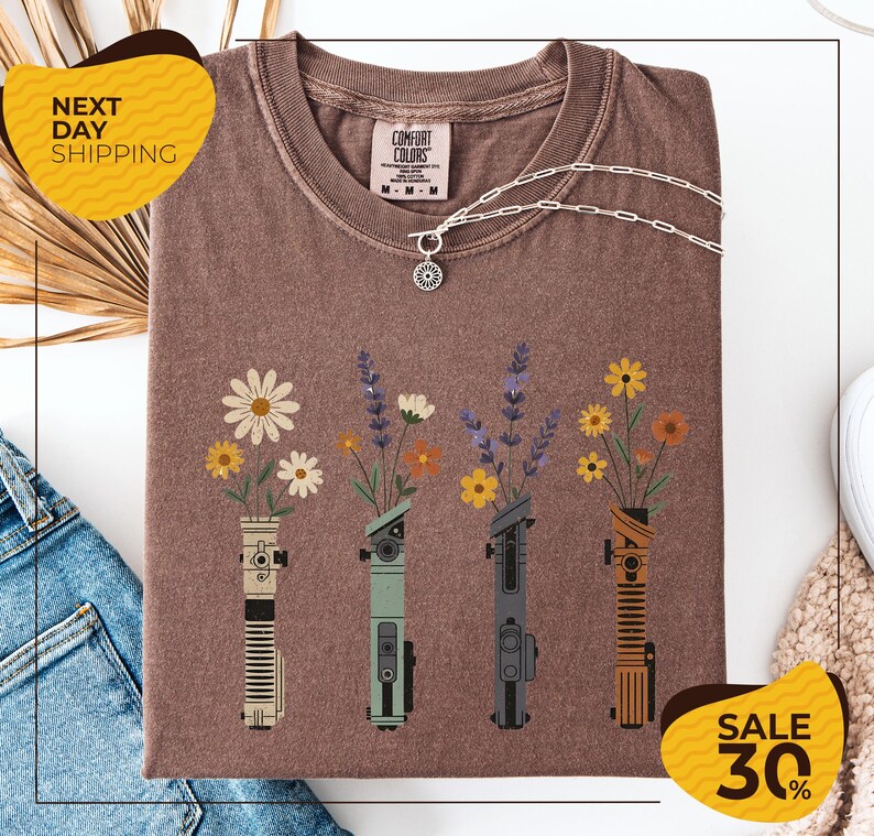 a brown t-shirt with a graphic design of four different colored vases holding flowers, arranged in a row. The vases are depicted in various colors, including green, gray, orange, and yellow. The design also includes a pendant necklace and a chain. The t-shirt is displayed on a white background, and there are yellow and black sale signs indicating a 30% discount.