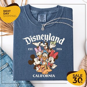 A blue t-shirt with a Disney-themed graphic, including Mickey Mouse, Minnie Mouse, Donald Duck, and Goofy, along with the text "Disneyland EST. 1955 CALIFORNIA". The t-shirt is displayed on a white background, and there are yellow and black sale signs indicating a 30% discount.