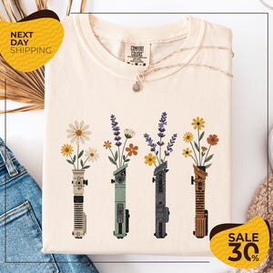a white t-shirt with a graphic design of four colorful vases holding flowers, arranged in a row. The vases are of different colors and shapes, creating a visually appealing pattern. The t-shirt is displayed on a white background, and there are some jeans and a sweater visible in the background. The image also includes text that reads "Next Day Shipping" and "SALE 30%", indicating that the t-shirt is available for immediate shipping and is on sale at a discounted price.