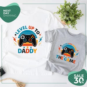 May include: A white t-shirt with a graphic of a black video game controller and the text "LEVEL UP TO DADDY" and a grey t-shirt with a black video game controller graphic and the text "PLAYER 2 HAS ENTERED THE GAME" with colorful accents.
