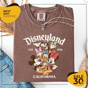 a brown t-shirt with a Disney-themed graphic, including Mickey Mouse, Minnie Mouse, Donald Duck, and Goofy, along with the text "Disneyland EST. 1955 CALIFORNIA". The t-shirt is displayed on a white background, and there are yellow and black sale signs indicating a 30% discount.