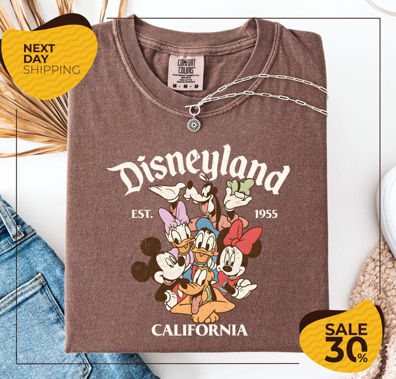 a brown t-shirt with a Disney-themed graphic, including Mickey Mouse, Minnie Mouse, Donald Duck, and Goofy, along with the text "Disneyland EST. 1955 CALIFORNIA". The t-shirt is displayed on a white background, and there are yellow and black sale signs indicating a 30% discount.