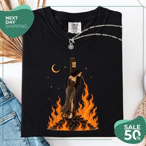 May include: A black t-shirt featuring a graphic print of a person with long dark hair tied to a wooden post, surrounded by bright orange flames. The design includes a crescent moon and small stars in the background. A silver chain necklace rests on the collar.