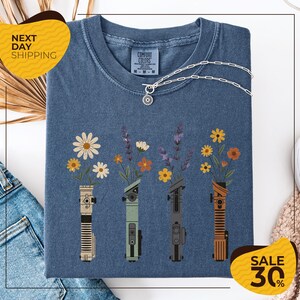 a blue t-shirt with a graphic design of four different colored vases holding flowers, arranged in a row. The vases are depicted in various colors, including blue, green, orange, and brown. The design also includes a small charm hanging from the vase. The t-shirt is displayed on a white background, and there are some jeans and a sweater visible in the background.