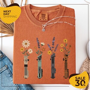 a t-shirt with a floral design, including four vases with flowers and various other objects, such as a necklace and a pair of jeans. The t-shirt is displayed on a white background, and there are yellow and black sale signs indicating a 30% discount.