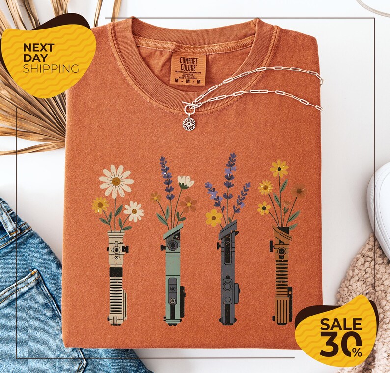 a t-shirt with a floral design, including four vases with flowers and various other objects, such as a necklace and a pair of jeans. The t-shirt is displayed on a white background, and there are yellow and black sale signs indicating a 30% discount.