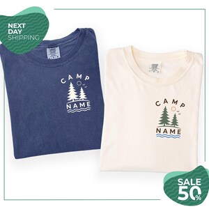 two t-shirts with the same design, one blue and one white, both featuring the text "CAMP NAME" and a graphic of trees. The t-shirts are folded and placed side by side, with a green and white sale banner in the background.