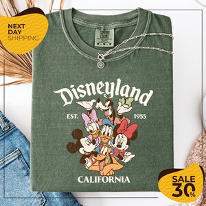a green t-shirt with a Disney-themed graphic, including Mickey Mouse, Minnie Mouse, Donald Duck, and Goofy, along with the text "Disneyland EST. 1955 CALIFORNIA". The t-shirt is displayed on a white background, and there are yellow and black sale signs indicating a 30% discount.