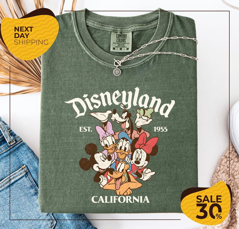 a green t-shirt with a Disney-themed graphic, including Mickey Mouse, Minnie Mouse, Donald Duck, and Goofy, along with the text "Disneyland EST. 1955 CALIFORNIA". The t-shirt is displayed on a white background, and there are yellow and black sale signs indicating a 30% discount.