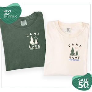 two t-shirts with the same design, one green and one white, both featuring the text "CAMP NAME" and a graphic of trees and waves. The t-shirts are folded and placed side by side, with a green and white background.
