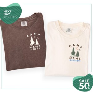 two t-shirts with the same design, one brown and one white, both featuring the text "CAMP NAME" and a graphic of trees. The brown t-shirt is folded, while the white t-shirt is unfolded. The image also includes text that reads "SALE 50%" and "NEXT DAY SHIPPING", suggesting that the t-shirts are on sale and can be ordered for the next day.