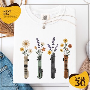 a white t-shirt with a graphic design of four colorful vases holding flowers, arranged in a row. The vases are of different colors and shapes, creating a visually appealing pattern. The t-shirt is displayed on a white background, and there are yellow and brown accents in the image.