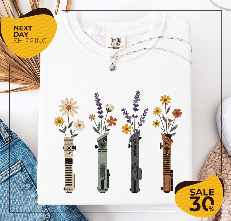 a white t-shirt with a graphic design of four colorful vases holding flowers, arranged in a row. The vases are of different colors and shapes, creating a visually appealing pattern. The t-shirt is displayed on a white background, and there are yellow and brown accents in the image.