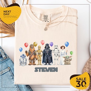 a t-shirt with a graphic design of various Star Wars characters, including Chewbacca, Darth Vader, Stormtrooper, and Yoda, along with balloons.