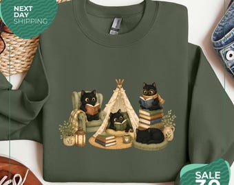 Könnte beinhalten: Ein olivgrünes Sweatshirt mit Rundhalsausschnitt und einer grafischen Illustration von fünf schwarzen Katzen in einer gemütlichen Umgebung. Die Katzen sind beim Lesen von Büchern dargestellt, sitzen in einem Sessel, in einem Tipi und auf Büchern.
