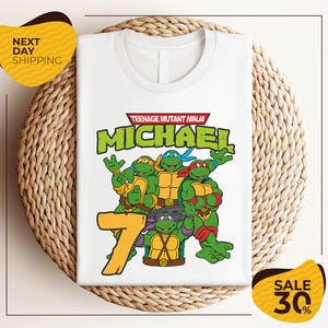May include: White t-shirt featuring the Teenage Mutant Ninja Turtles. The shirt has a graphic of the four turtles, the name "MICHAEL", and the number "7" in a yellow font. The shirt is displayed on a woven surface.
