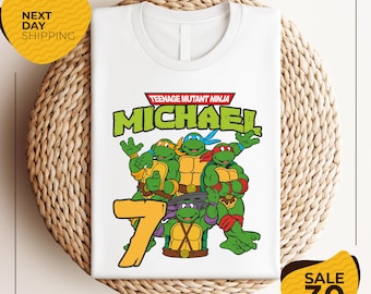 Könnte beinhalten: Weißes T-Shirt mit den Teenage Mutant Ninja Turtles. Das Shirt hat eine Grafik der vier Turtles, den Namen "MICHAEL" und die Zahl "7" in gelber Schrift. Das Shirt wird auf einer gewebten Oberfläche präsentiert.