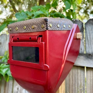 May include: A red welding helmet with a snakeskin-patterned top and brass rivets. The helmet has a dark viewing lens and is mounted on a wooden post. The background features a wooden fence and green foliage.