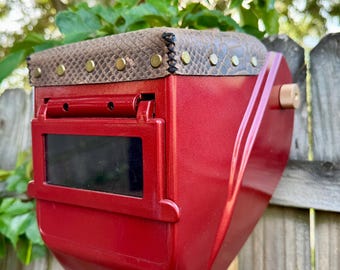 May include: A red welding helmet with a snakeskin-patterned top and brass rivets. The helmet has a dark viewing lens and is mounted on a wooden post. The background features a wooden fence and green foliage.