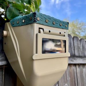 Puede incluir: Un casco de soldadura beige con una parte superior verde texturizada, asegurada con remaches de color latón. El casco tiene una ventana de visualización rectangular y está montado en un poste de madera contra una valla de madera y un cielo azul.
