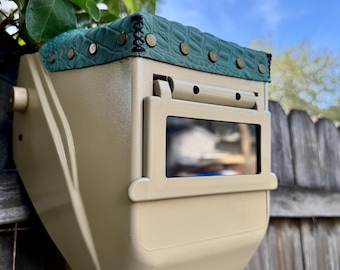 May include: A tan welding helmet with a green, textured top, secured with brass-coloured rivets. The helmet has a rectangular viewing window and is mounted on a wooden post against a wooden fence and a blue sky background.