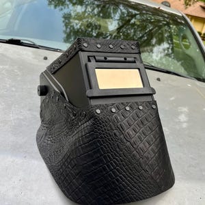 Pode incluir: Capacete de soldagem preto com um padr&atilde;o texturizado semelhante &agrave; pele de crocodilo na frente e nas laterais. O capacete tem uma janela de visualiza&ccedil;&atilde;o retangular e &eacute; adornado com rebites prateados. A parte superior tamb&eacute;m &eacute; preta e tem rebites prateados.