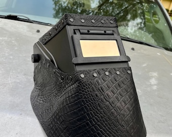 May include: Black welding helmet with a crocodile-effect textured finish. The helmet features a rectangular viewing window and is detailed with silver rivets. The top is also black and has silver rivets.