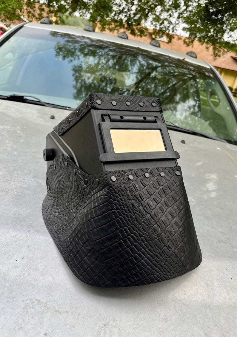 Pode incluir: Capacete de soldagem preto com um padr&atilde;o texturizado semelhante &agrave; pele de crocodilo na frente e nas laterais. O capacete tem uma janela de visualiza&ccedil;&atilde;o retangular e &eacute; adornado com rebites prateados. A parte superior tamb&eacute;m &eacute; preta e tem rebites prateados.